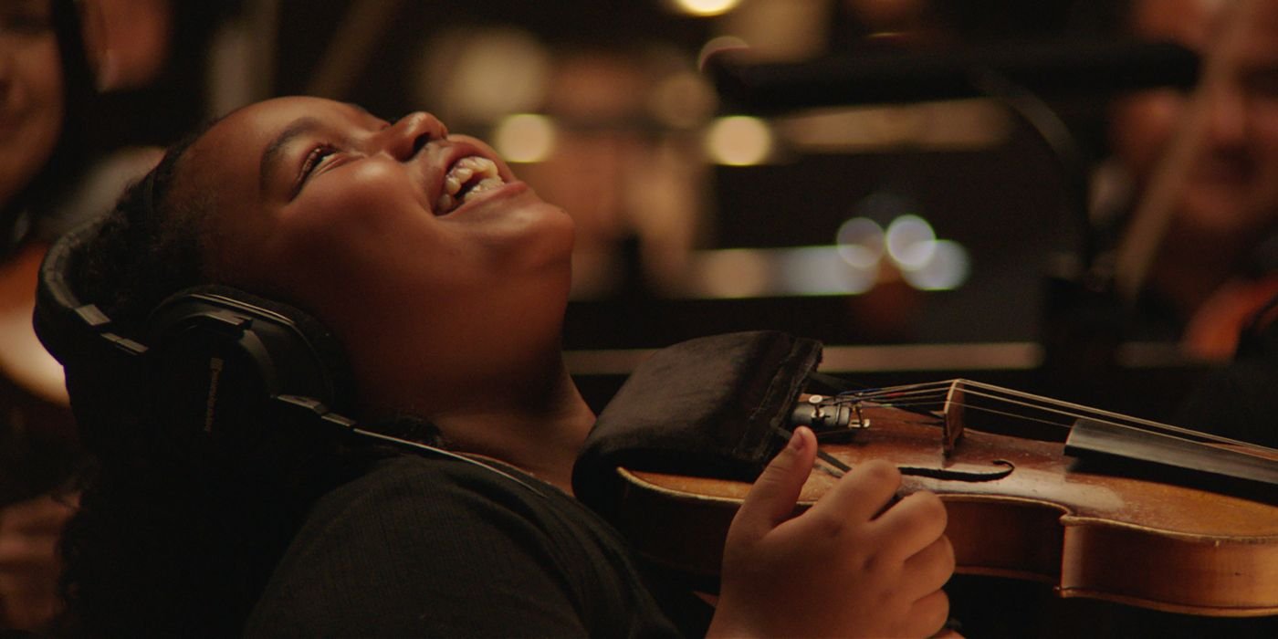 A young girl wearing headphones and playing the violin to an audience in The Last Repair Shop
