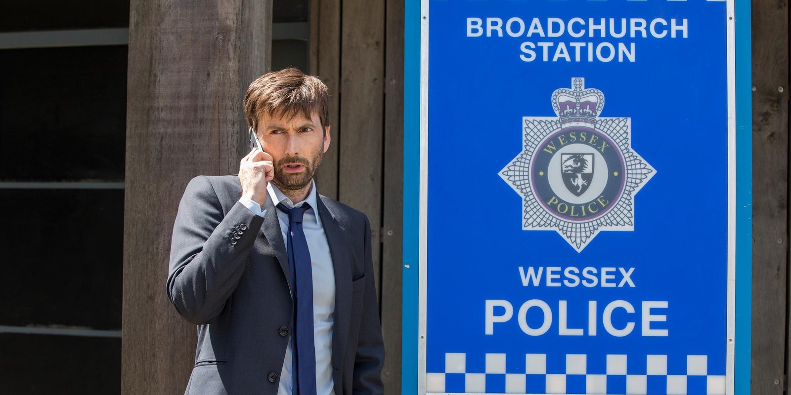 David Tennant as Alec Hardy looking serious, while standing outside the police station on the phone, in Broadchurch.