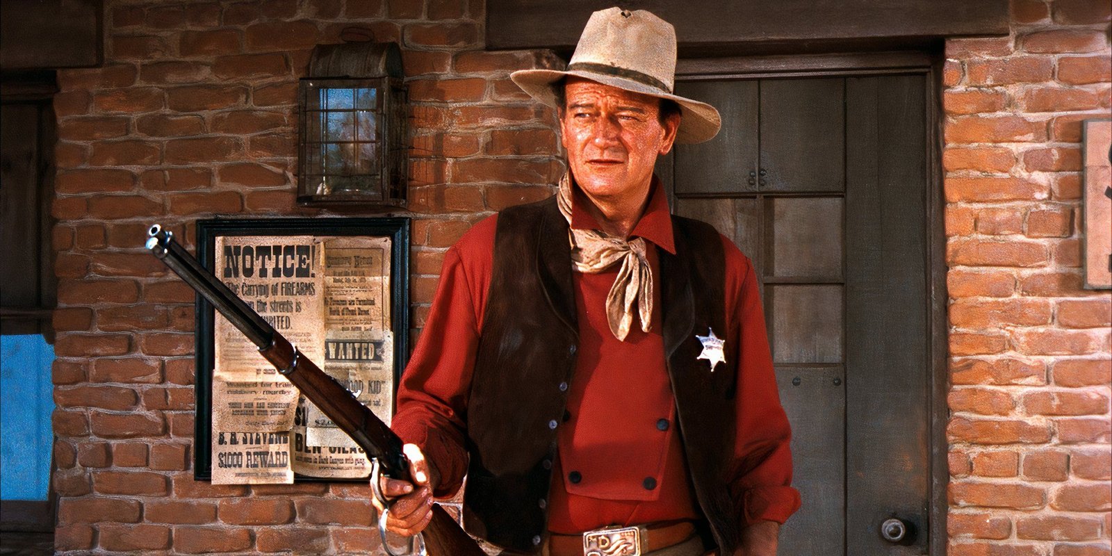 What John Wayne's Real Name Was & Why He Changed It 4 John Wayne holds up a rifle while standing in front of the Sheriff's Office in Rio Bravo