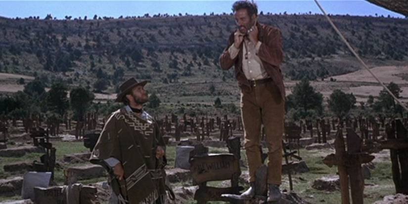 A still from The Good, The Bad, And The Ugly. Blondie holds Tuco at gunpoint while he sets up a noose for himself. There is a large cemetery in the background.